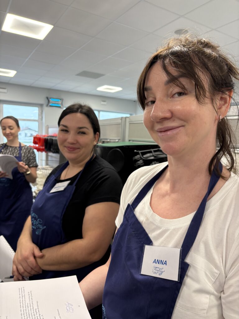 Dwie kobiety w fartuchach cukierniczych stoją w profesjonalnej pracowni i uśmiechają się do zdjęcia.