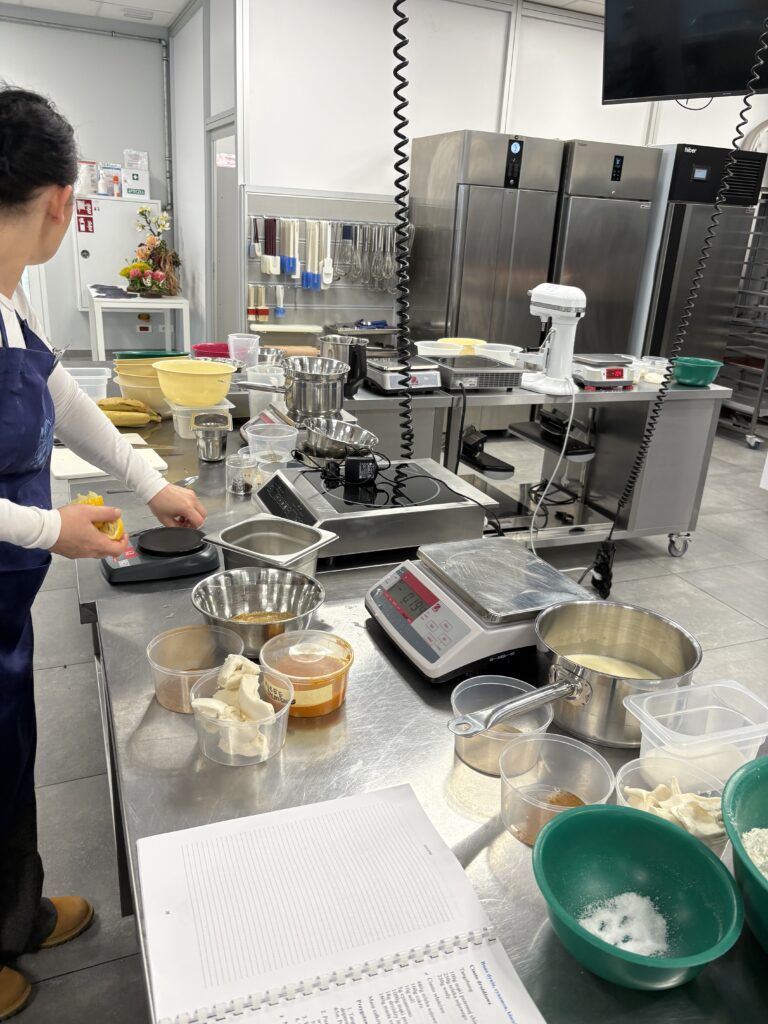 Osoba waży składniki na wadze kuchennej przy stanowisku roboczym z przygotowanymi produktami.