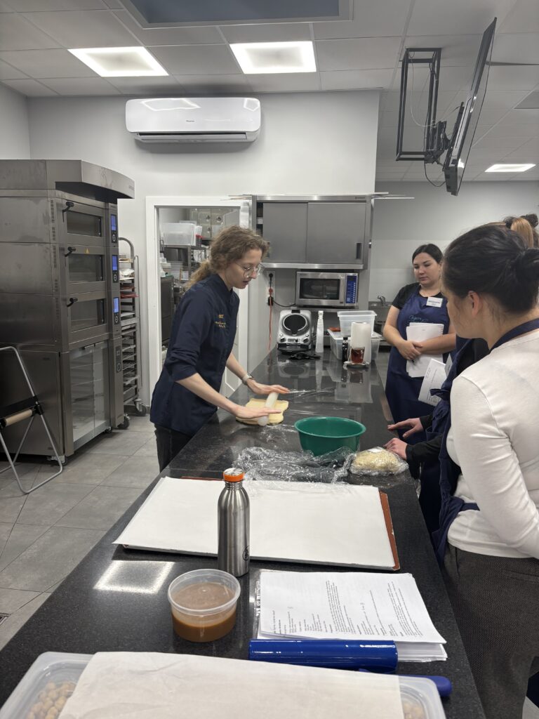 Instruktorka omawia proces przygotowania ciasta przy stole roboczym z uczestniczkami szkolenia.