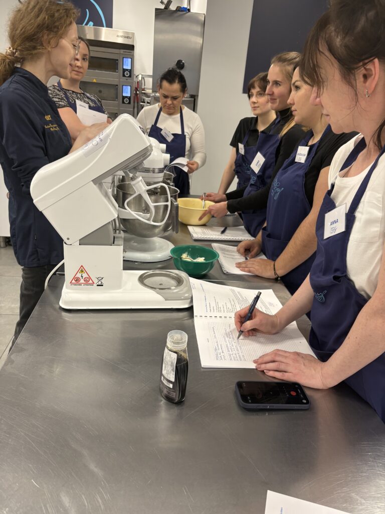 Grupa kobiet w fartuchach obserwuje pokaz użycia miksera planetarnego w pracowni cukierniczej.