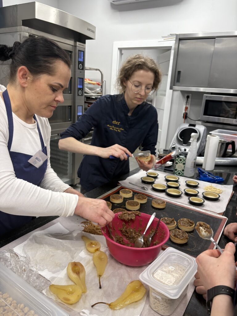 Uczestniczki szkolenia dekorują małe tarty nadzieniem owocowym w pracowni cukierniczej.
